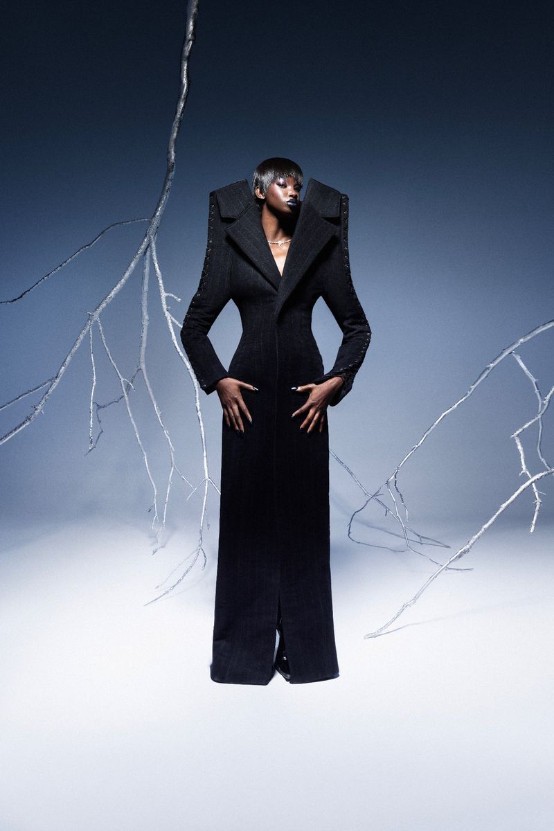 An African American woman stands in a formal black coat dress with architectural shoulders against a backdrop with silver branches and fog.
