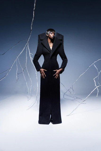 An African American woman stands in a formal black coat dress with architectural shoulders against a backdrop with silver branches and fog.