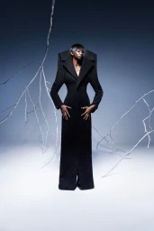 A Black female model stands wearing a dramatic, exaggerated black sculptural coat framed by bare white branches in a studio setting.