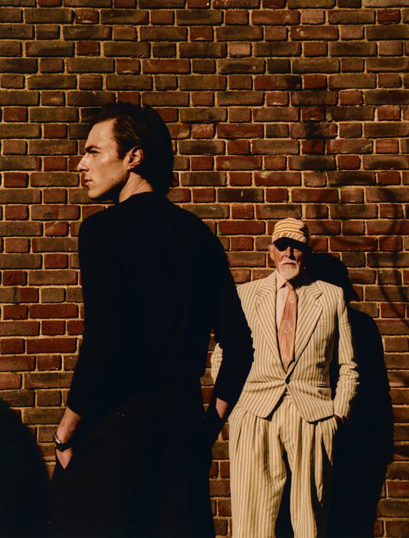 Two men in contrasting black knitwear and a striped tailored suit posing in front of a brick wall in harsh sunlight.