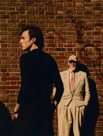 Two men in contrasting black knitwear and a striped tailored suit posing in front of a brick wall in harsh sunlight.