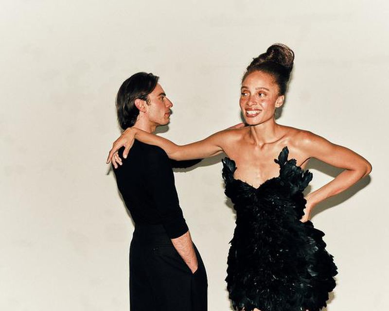 Portrait of a smiling woman in a feathered black dress, arm on the shoulder of a man in black, against a neutral backdrop.