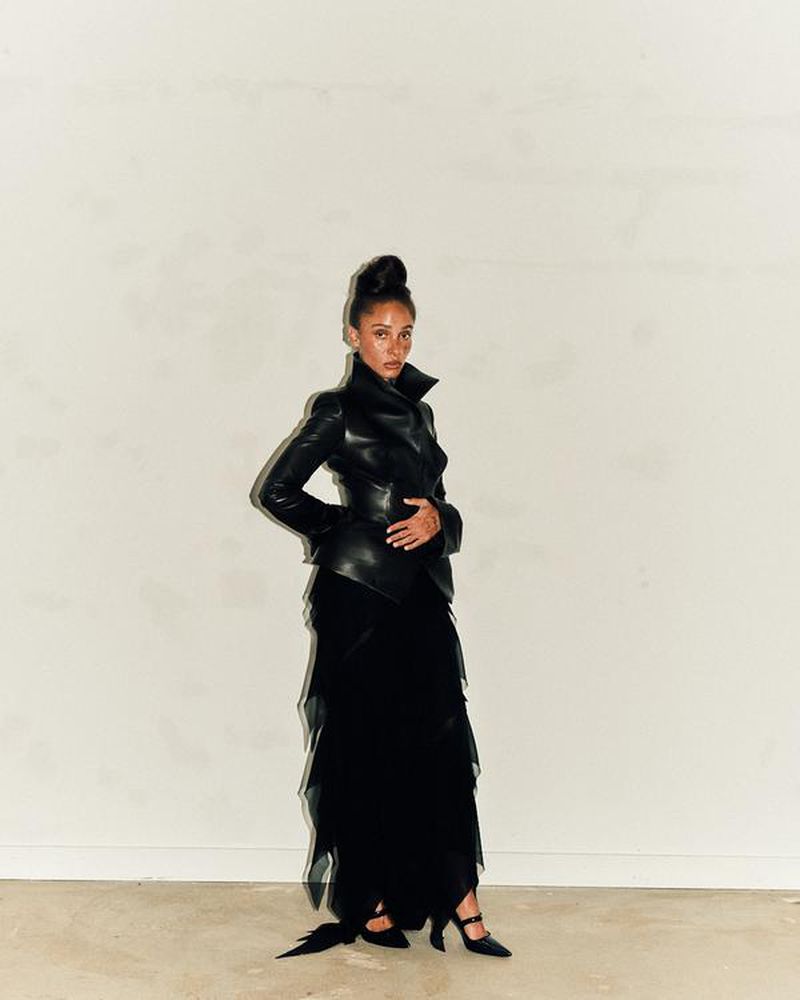 Woman in elegant black leather jacket and layered tulle dress strikes a pose against a neutral backdrop in a fashion-forward photograph.