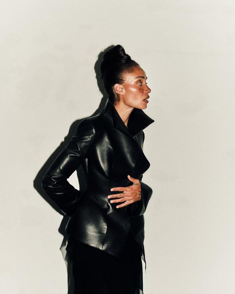Model Adwoa Aboah wearing a structured black leather jacket with an updo hairstyle against a light grey studio background.