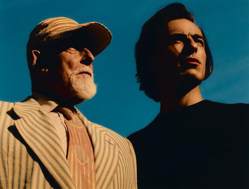 Low angle portrait of an elderly bearded man in a striped suit and cap beside a younger man against a clear blue sky.