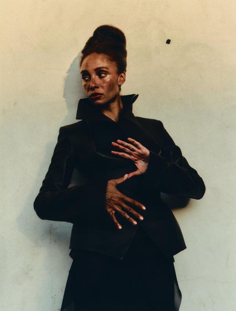 A striking editorial fashion portrait of a Black female model in a dark jacket against a textured neutral wall, with hands gently posed.