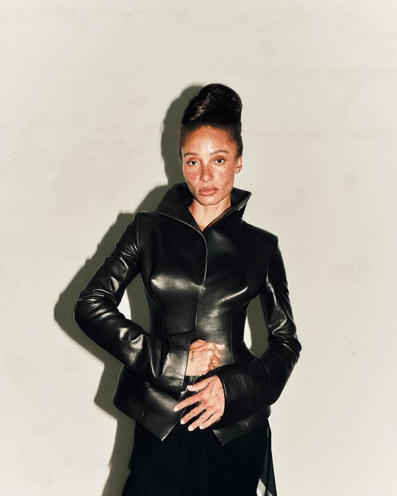 A Black woman with an elegant updo poses indoors wearing a black leather jacket with structured shoulders against a neutral background.