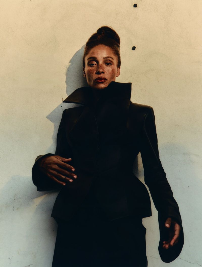 Elegant portrait of a Black woman in a high-collared black jacket against a textured beige wall, highlighting fashion and refined style.