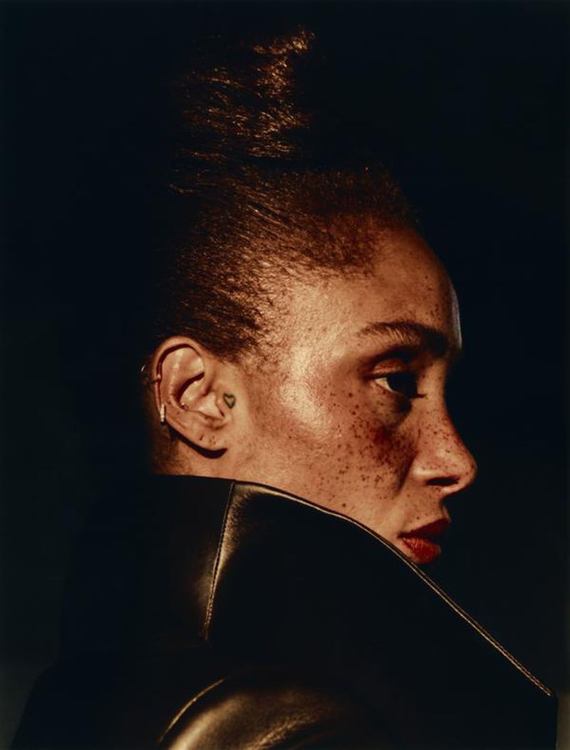 Striking close-up portrait of a woman with freckles and her hair in a high bun, wearing a dark leather jacket against a dark background.