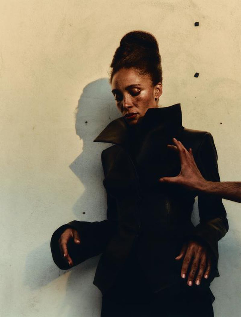 African-American woman in dark jacket poses indoors with hand on chest in studio lighting, showcasing fashion and beauty.