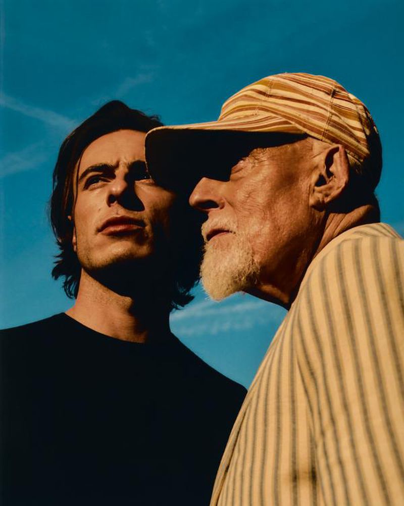 An eye-level outdoor shot features a young adult male and an older adult male in close proximity against a clear blue sky.