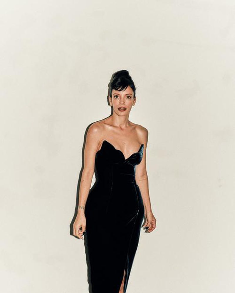 A full shot of a woman in a strapless black velvet dress with a thigh-high slit, posing against a neutral background in portrait orientation.