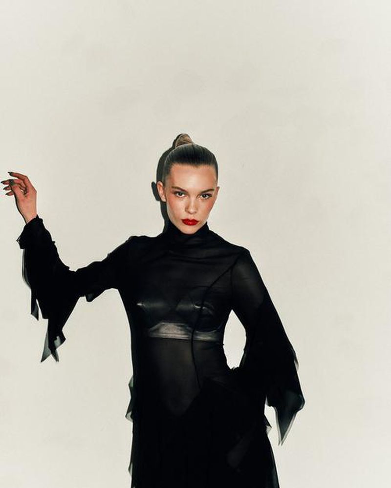 Fashionable woman in a sheer black dress with sleeves, striking red lipstick, and a sleek ponytail poses against a stark white background.