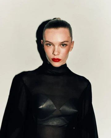 Headshot of a fair-skinned adult woman with red lipstick, slicked-back hair, and a black mesh top in a studio setting.