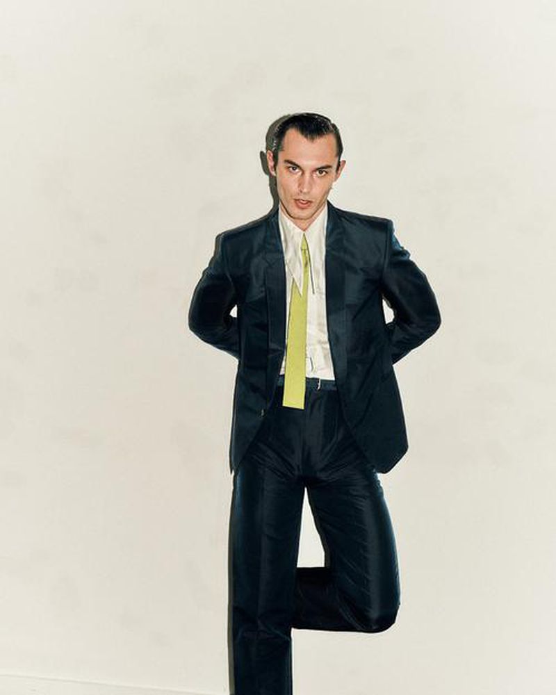 Male model poses in a dark suit with a pale green tie against a neutral background. He's leaning slightly to the side.
