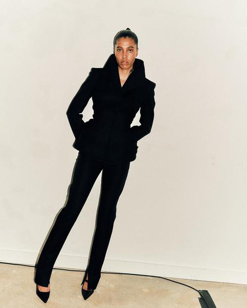 Woman in a black tailored suit stands against a pale background in a fashion pose, showcasing sleek, modern design and elegance.