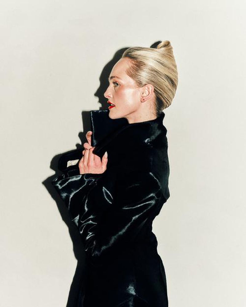 Studio portrait of an adult woman with blonde hair in a black coat, striking a pose against a neutral backdrop. Fashion and beauty theme.