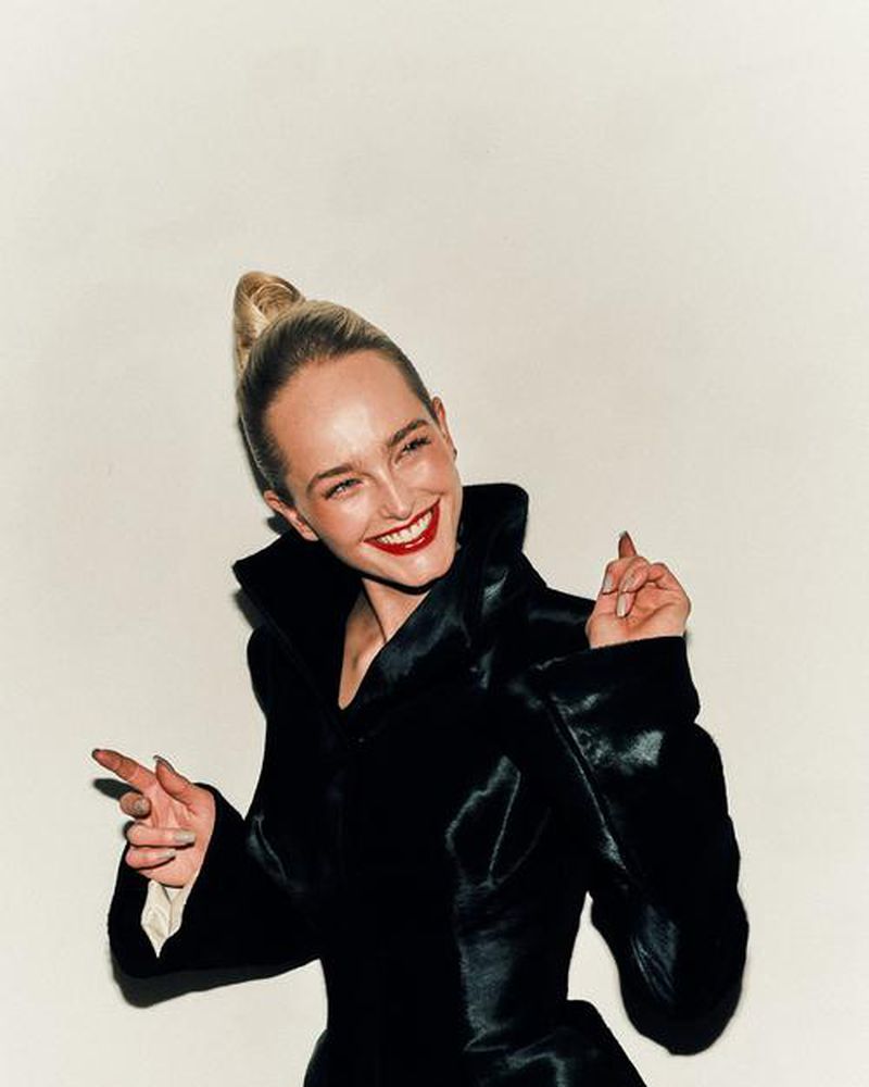 Blonde woman in a dark coat with red lipstick poses in a studio, smiling and pointing at the camera against a white backdrop.