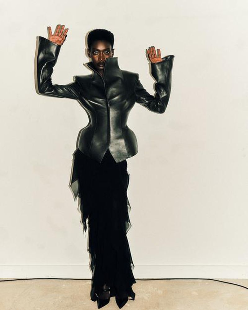 A striking fashion portrait of a black female model with hands raised, wearing a sculptural black leather jacket and layered black skirt.
