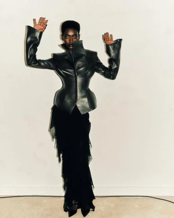 A striking fashion portrait of a black female model with hands raised, wearing a sculptural black leather jacket and layered black skirt.