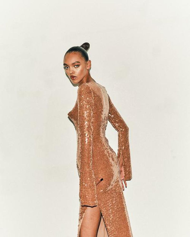 Elegant adult female in a long-sleeved, tan sequin dress with a thigh-high slit posing in a studio against a neutral background.
