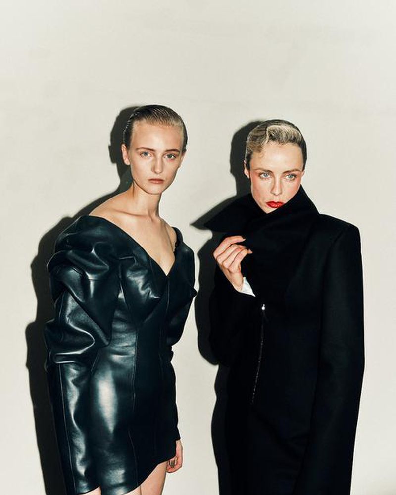 Two adult women in black dresses pose indoors against a white wall, highlighting fashion and style in a studio-like environment.