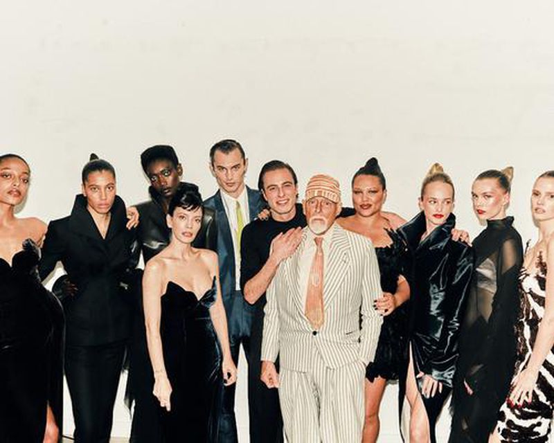 A group of diverse fashion models pose in an indoor photo studio, wearing various black and patterned outfits, with a man in a striped suit.