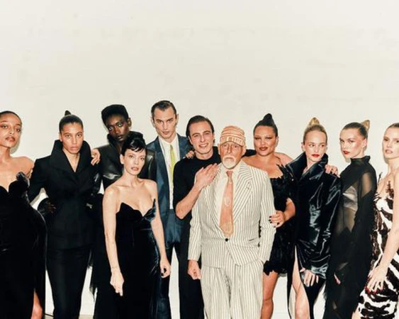 A group of diverse fashion models pose in an indoor photo studio, wearing various black and patterned outfits, with a man in a striped suit.