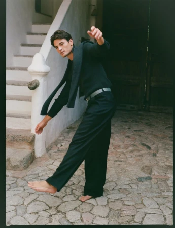 Barefoot male model in a dark sweater and trousers balances on a stone street next to white stucco stairs and a dark opening.