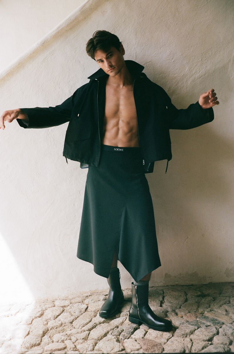 Fashion shot of a muscular man posing with open jacket and skirt against a white wall, showcasing modern style and design.