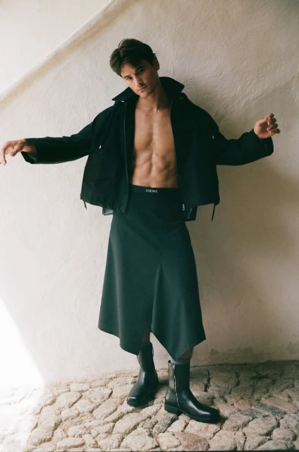 Fashion shot of a muscular man posing with open jacket and skirt against a white wall, showcasing modern style and design.