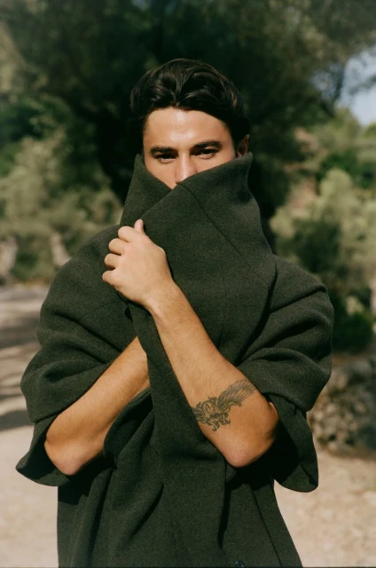 Portrait of a man with dark hair wearing a dark green coat covering his face, arms crossed, and standing outdoors in a natural setting.