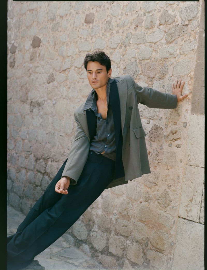Portrait of a young adult man in stylish casual attire leaning against a stone wall, captured in natural light with a medium shot.
