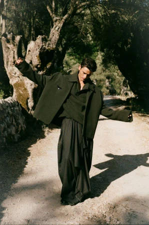A stylish adult male poses on a sunlit path outdoors, wearing a dark jacket, shirt, and wide-legged pants, surrounded by trees and natural light.