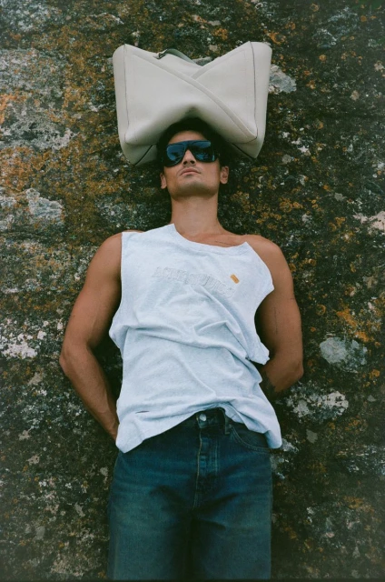 A man in sunglasses and denim lying on a rock with a geometric tote bag balancing on his head in an outdoor setting.