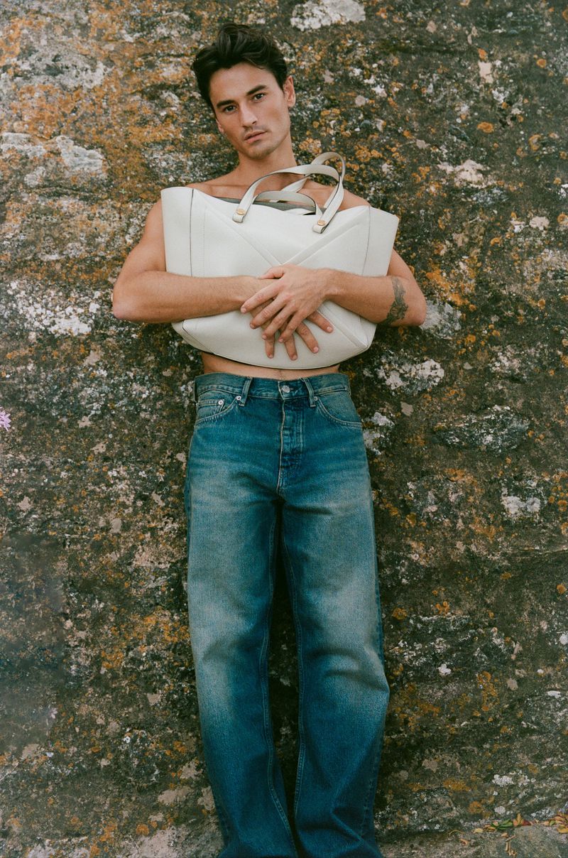 Shirtless man in blue denim jeans holding a large cream leather tote bag against a lichen-covered stone wall.