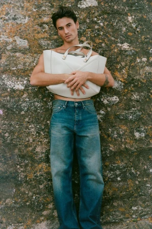 Shirtless man in blue denim jeans holding a large cream leather tote bag against a lichen-covered stone wall.