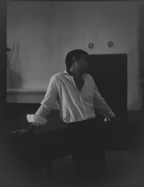 Monochrome portrait of an adult male in unbuttoned white shirt and black trousers, indoors with an expressive pose and soft lighting.