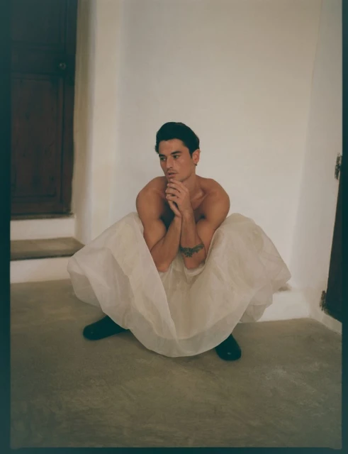 Shirtless man in a white tulle skirt sits with his hands together in an indoor space with a door in the background.
