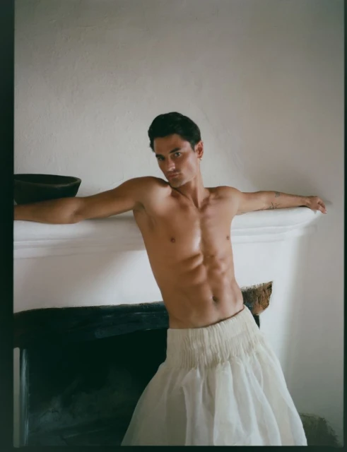 Portrait of a muscular adult male leaning against a fireplace, wearing a sheer white ruffled skirt in an indoor setting.