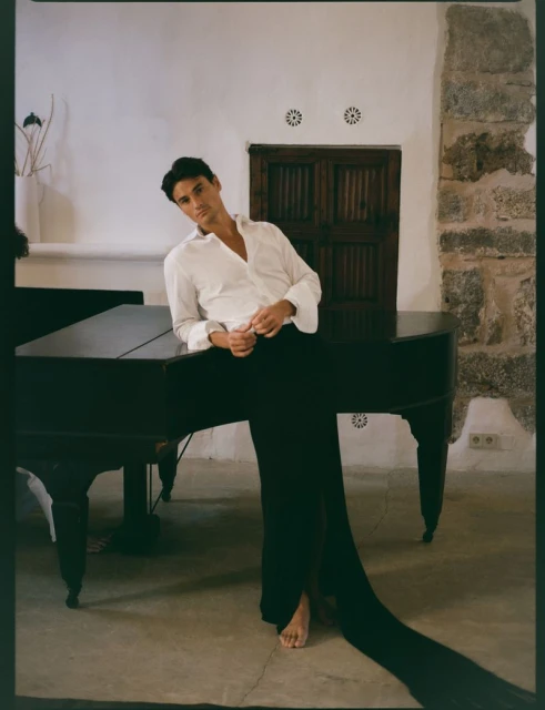 Young man poses barefoot in a white shirt and black pants near a piano indoors against a textured wall, creating a classic portrait.