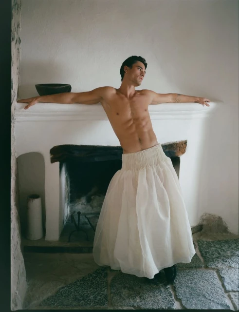 Shirtless man in a white skirt leans against a fireplace mantel in a rustic stone room, creating a visually striking, high-fashion image.