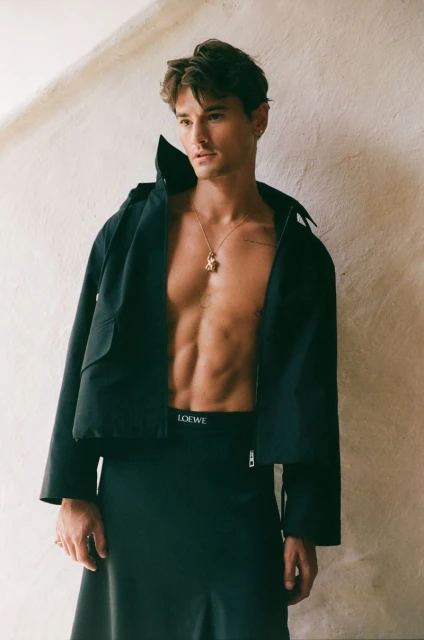 Man poses in jacket and skirt against neutral background. Open jacket reveals torso. Gold necklace visible.