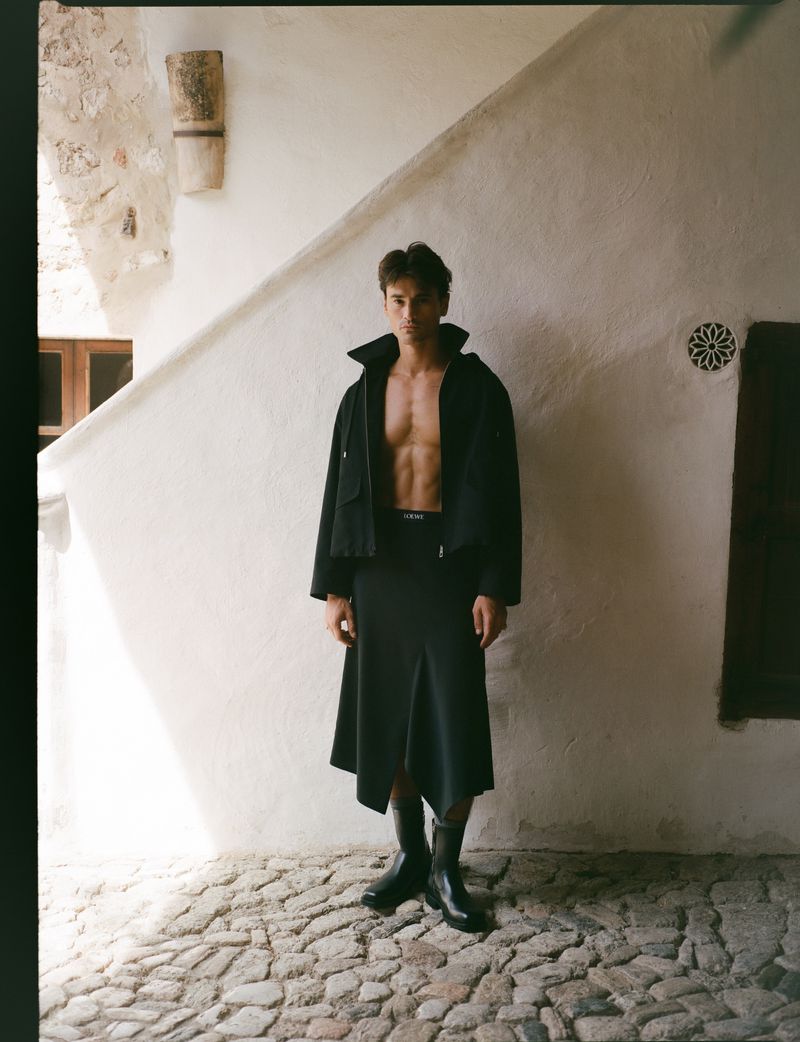 A man in a dark skirt and jacket strikes a pose against a white wall with a stone pathway. He is shirtless in the indoor setting.