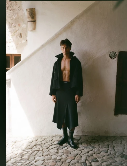 A man in a dark skirt and jacket strikes a pose against a white wall with a stone pathway. He is shirtless in the indoor setting.