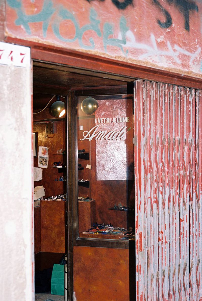 Exterior view of a vintage shop with a partially open shutter, revealing shelves of glasses and a retro aesthetic in an urban setting.
