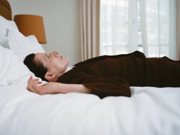 Adult female lying on a white bed in a light-filled room with a window in the background; wearing a brown robe and facing to the side.