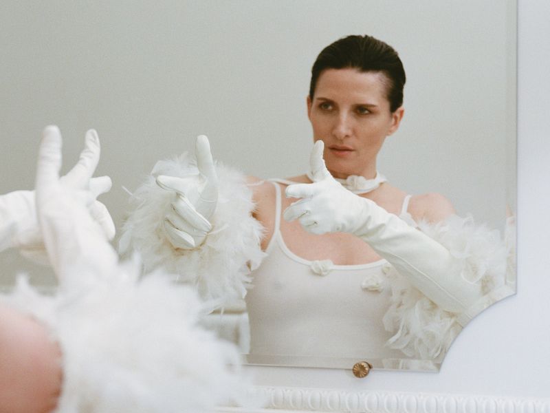 Adult woman in white gloves and top poses with a gun-finger gesture, reflected in a mirror against a neutral background.