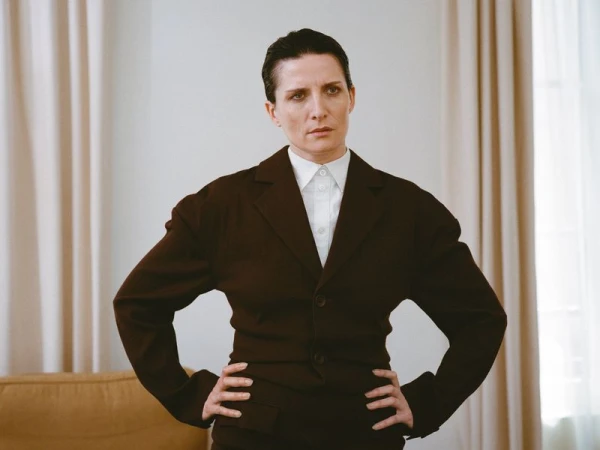 Portrait of a woman with short dark hair wearing a brown suit and white collared shirt posing indoors with hands on her hips.