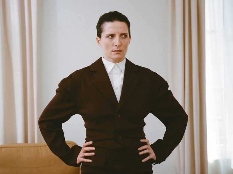 Serious woman in brown suit with hands on hips stands in front of white curtains and light yellow couch indoors, looking to the side.
